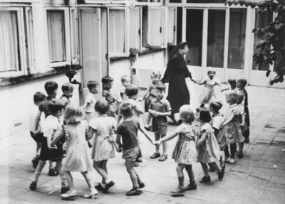 876951 Afbeelding van een groep kleuters die met een Zuster Augustines die in haar functie als 'kleuterjuf' een ...